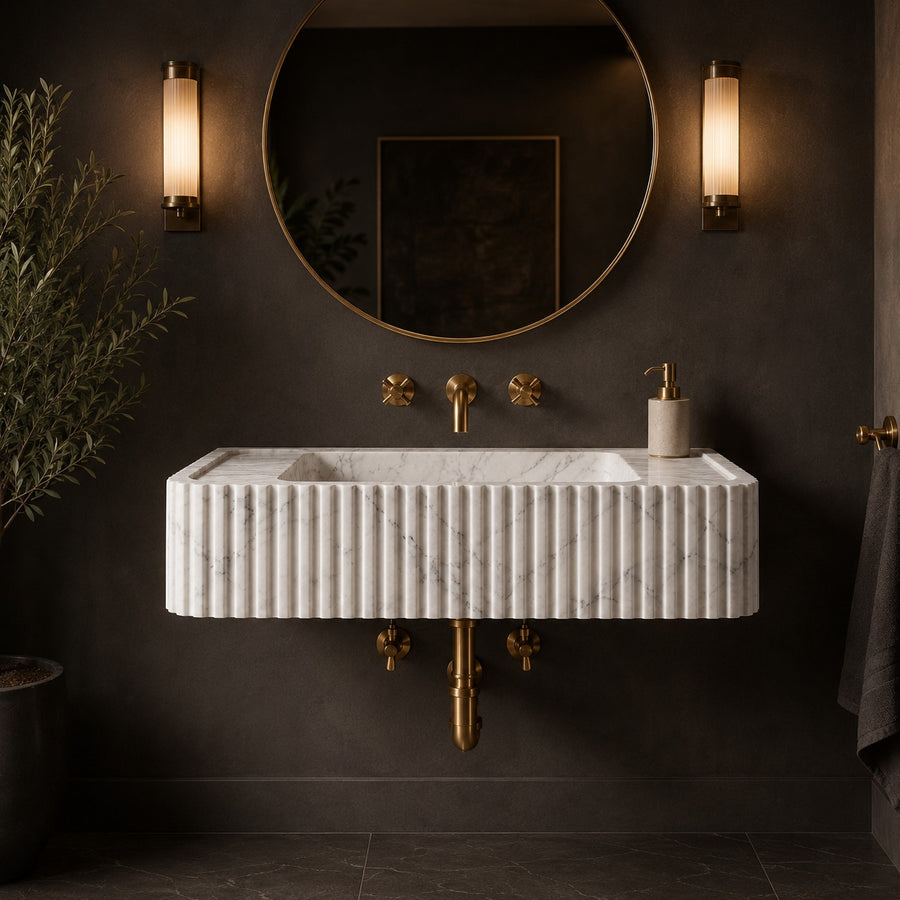 A modern wall-mounted bathroom sink with a ribbed white marble front and gold hardware, installed against a dark charcoal wall beneath a round mirror and between two vertical sconces.