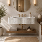 A modern floating bathroom vanity with a ribbed white marble sink, gold faucets, a wooden shelf, and a round wall mirror in a softly lit neutral bathroom setting.
