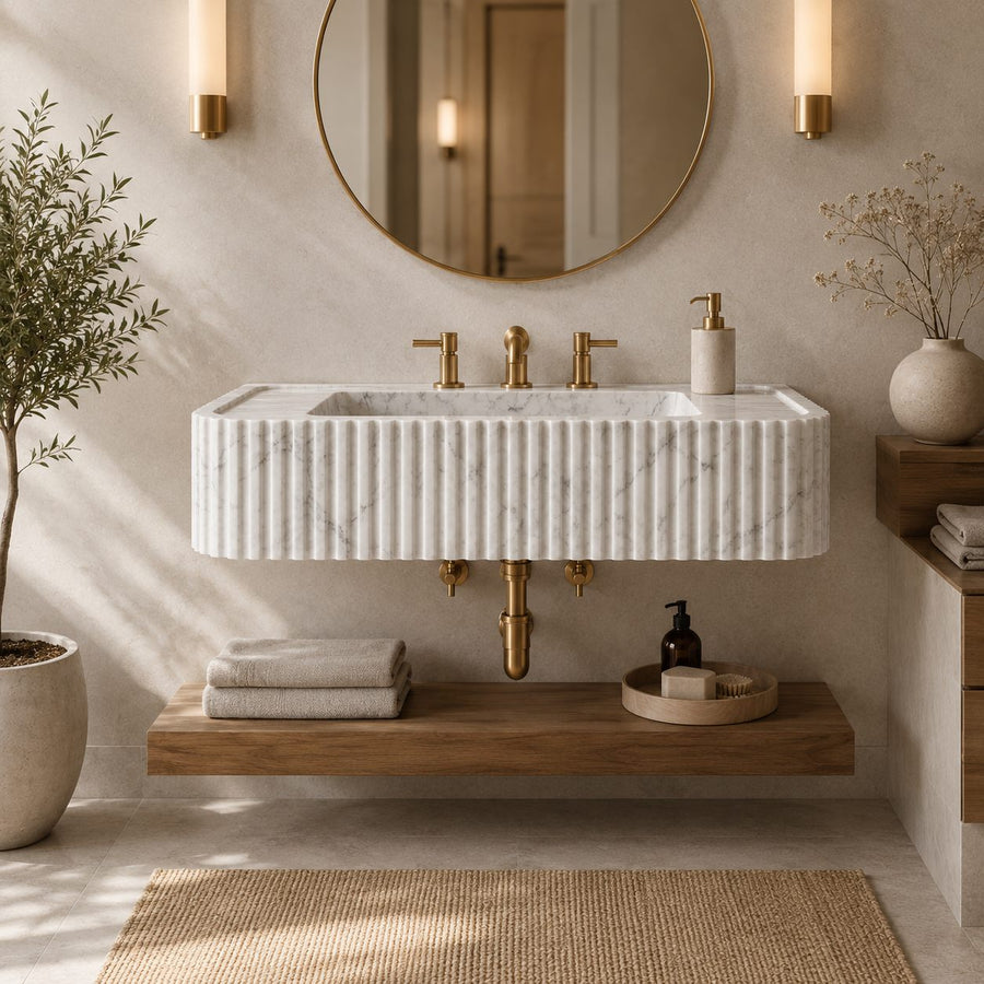 A modern floating bathroom vanity with a ribbed white marble sink, gold faucets, a wooden shelf, and a round wall mirror in a softly lit neutral bathroom setting.