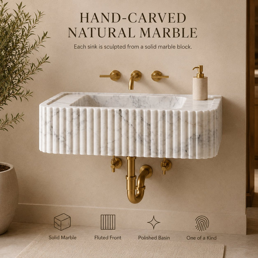 A wall-mounted rectangular sink made of natural marble with a fluted front and polished basin, shown with brass faucet hardware and a small soap dispenser against a beige wall. Text above reads 'HAND-CARVED NATURAL MARBLE' and icons below highlight solid marble, fluted front, polished basin, and one-of-a-kind quality.