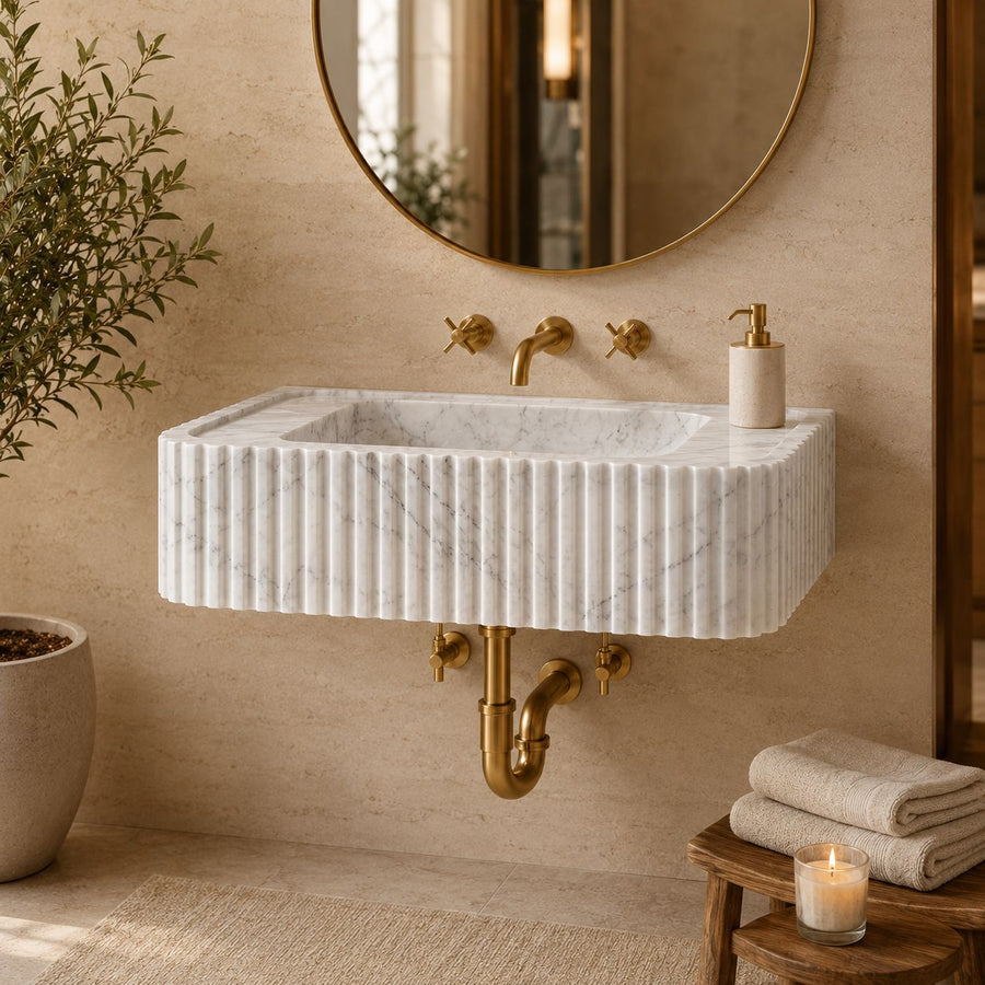 A modern wall-mounted rectangular sink made of white fluted marble with a brushed gold brass faucet and exposed drain, set in a warm beige bathroom with a round mirror, folded towels, and a lit candle.