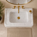 A white marble washbasin with a fluted edge and a recessed sink, paired with a brushed gold faucet, drain, and matching round mirror. A small potted plant sits on the left, and a beige bath mat is on the floor below.