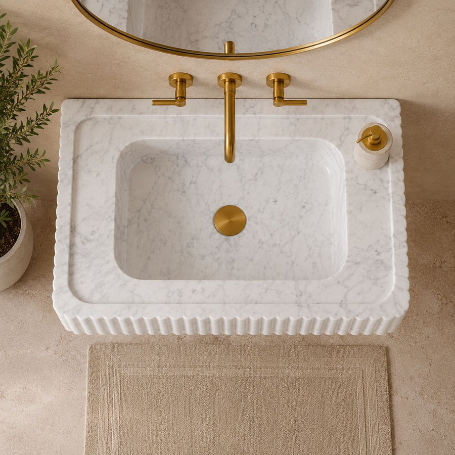 A white marble washbasin with a fluted edge and a recessed sink, paired with a brushed gold faucet, drain, and matching round mirror. A small potted plant sits on the left, and a beige bath mat is on the floor below.