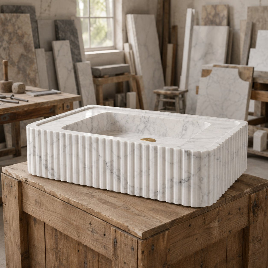 A rectangular white marble sink basin with fluted edges, displayed on a rustic wooden table inside a stone and wood workshop.