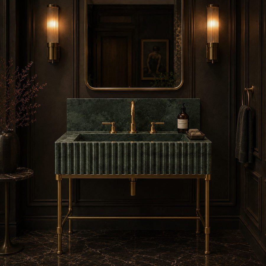 A luxury bathroom vanity featuring a dark green fluted marble sink and matching backsplash on a gold metal stand, set against dark wood paneled walls with wall sconces and a mirror.