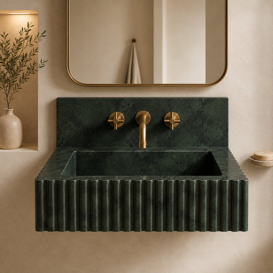 A floating rectangular sink made of dark green fluted stone with a matching backplate, paired with matte brass fixtures and a large rounded mirror on a warm beige wall.