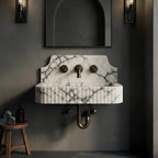 A modern wall-mounted bathroom sink with a white marble basin featuring dramatic dark gray veining, set against a dark gray textured wall. The sink has a fluted front, a central round faucet, two separate round side knobs, and a visible brass P-trumpet pipe.