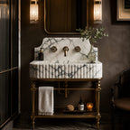 A luxury bathroom vanity featuring a white marble sink with dark veining and a fluted front, mounted on an ornate brass pedestal stand. The setup includes a matching marble backsplash, a central brass faucet, and a brass lower shelf holding a folded towel and toiletries, set against a dark brown wall with wall-mounted light fixtures.