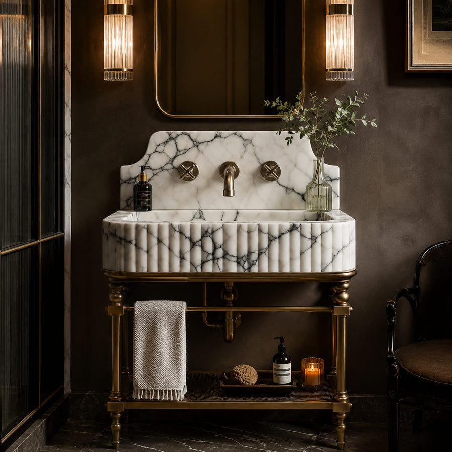 A luxury bathroom vanity featuring a white marble sink with dark veining and a fluted front, mounted on an ornate brass pedestal stand. The setup includes a matching marble backsplash, a central brass faucet, and a brass lower shelf holding a folded towel and toiletries, set against a dark brown wall with wall-mounted light fixtures.