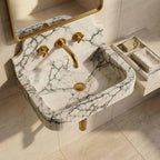 A wall-mounted bathroom sink with a white marble basin featuring dramatic black veining, paired with a brushed gold faucet, handles, and mirror frame. The sink is mounted on a beige tiled wall with a matching marble soap dish holding folded towels on the right.