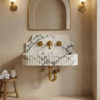 A wall-mounted marble sink with a white and green veined pattern, paired with a matching marble basin and gold-finished faucet, handles, and P-trumpet drain, set against a beige plaster wall with a gold-framed arch mirror and wall sconces.