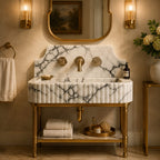 An elegant bathroom vanity featuring a white marble sink with dramatic black veining, a matching marble backplate, and a polished gold brass pedestal base and fixtures. The vanity is styled in a luxurious bathroom setting with a gold-framed mirror, wall sconces, a towel, and toiletries.