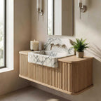 A modern floating bathroom vanity with a fluted light wood cabinet, a white marble sink basin, and a matching marble backsplash, styled in a warm beige bathroom with a mirror, sconces, a candle, and a potted fern.