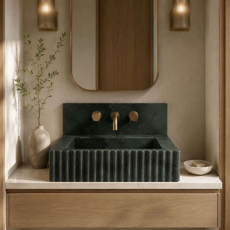 A modern bathroom vanity featuring a dark green ribbed stone sink with a matching backplate and brass faucet, set on a light marble countertop with a wooden cabinet below.