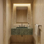 A modern bathroom vanity featuring a green marble sink with a fluted front, set against a warm beige travertine stone wall. A matching rectangular mirror is mounted above, with a black faucet and a white soap dispenser.