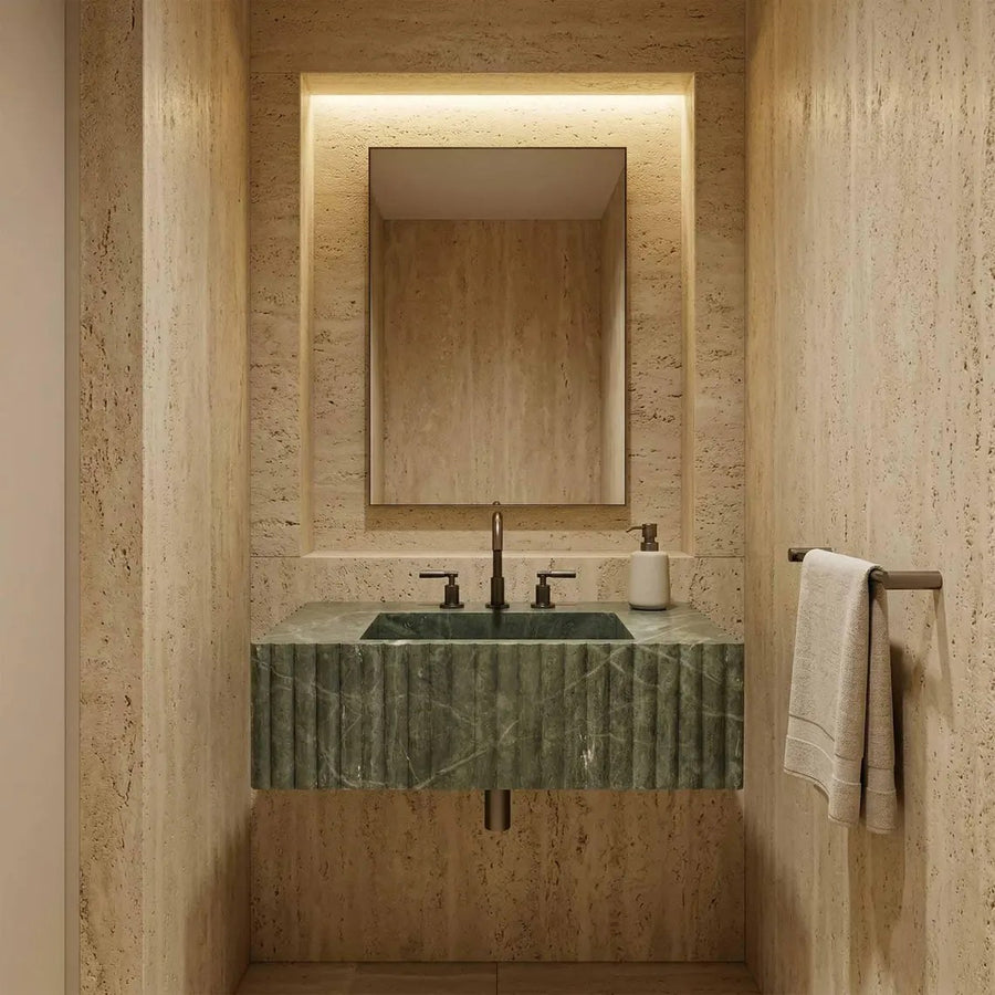 A modern bathroom vanity featuring a green marble sink with a fluted front, set against a warm beige travertine stone wall. A matching rectangular mirror is mounted above, with a black faucet and a white soap dispenser.