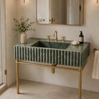 A modern bathroom vanity featuring a rectangular green marble sink with a fluted front, mounted on a gold metal stand. The sink is styled with a gold faucet, gold handles, a soap dish, a small vase with greenery, and a dark bottle, set against a warm beige wall with a gold-framed mirror.
