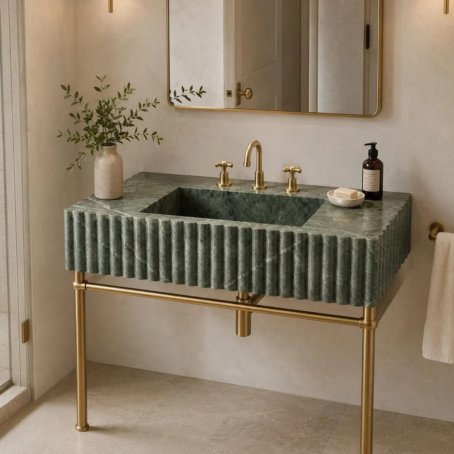 A modern bathroom vanity featuring a rectangular green marble sink with a fluted front, mounted on a gold metal stand. The sink is styled with a gold faucet, gold handles, a soap dish, a small vase with greenery, and a dark bottle, set against a warm beige wall with a gold-framed mirror.