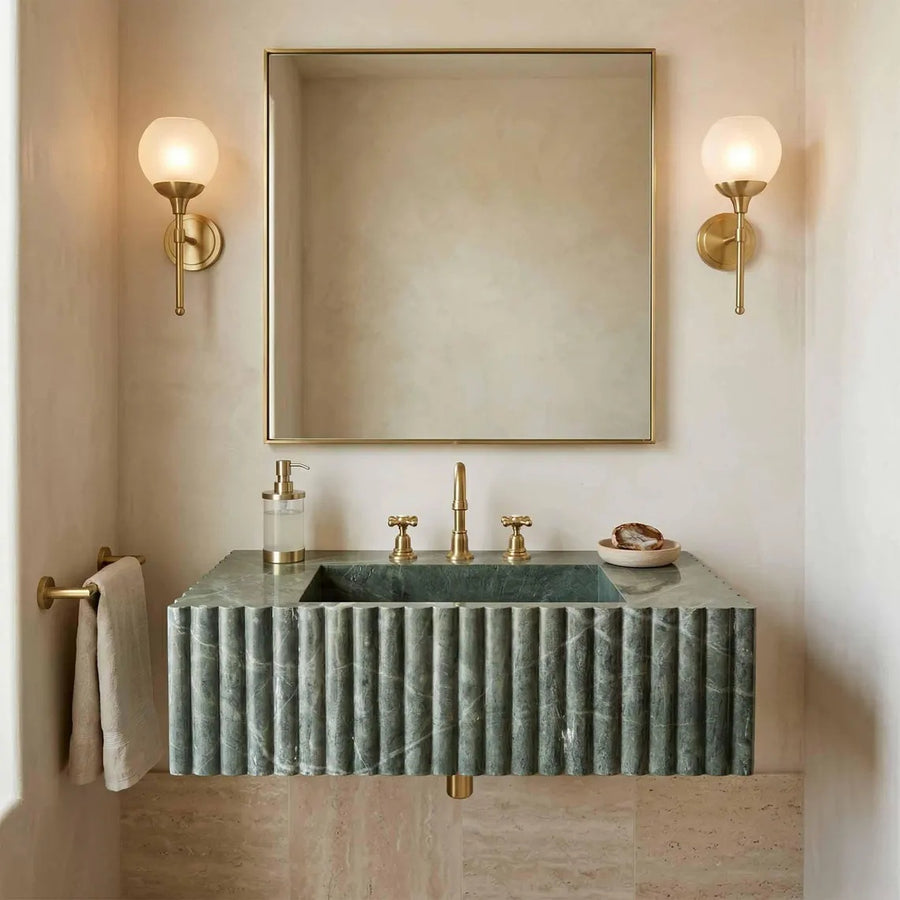 A modern bathroom vanity setup featuring a ribbed green marble sink with a matching marble countertop, a gold faucet, a large rectangular mirror, and two gold wall sconces on a beige wall.