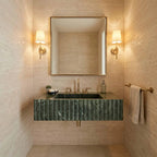 A modern bathroom vanity featuring a dark green marble sink with a fluted front, mounted against a beige stone wall. A large rectangular mirror with a gold frame hangs above, flanked by two warm wall sconces.
