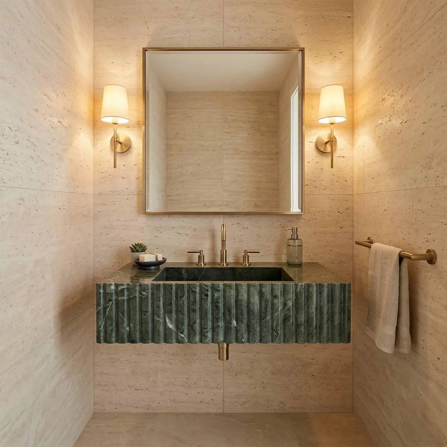 A modern bathroom vanity featuring a dark green marble sink with a fluted front, mounted against a beige stone wall. A large rectangular mirror with a gold frame hangs above, flanked by two warm wall sconces.