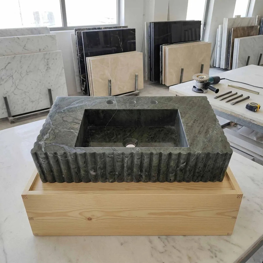A rectangular dark stone sink basin with a ribbed fluted edge, displayed in a light wood crate on a marble workbench in a showroom. Other marble slabs and construction tools are visible in the background.