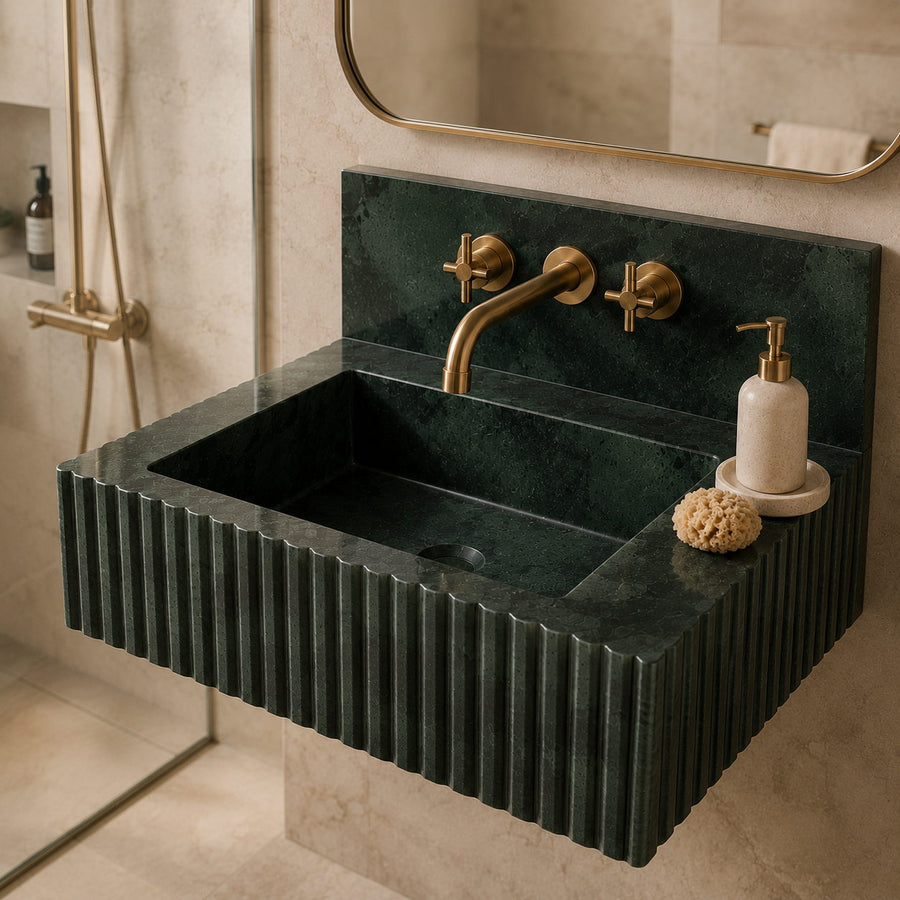 A floating rectangular sink made of dark green fluted stone, paired with a brushed brass wall-mounted faucet and a cream soap dispenser in a beige bathroom setting.