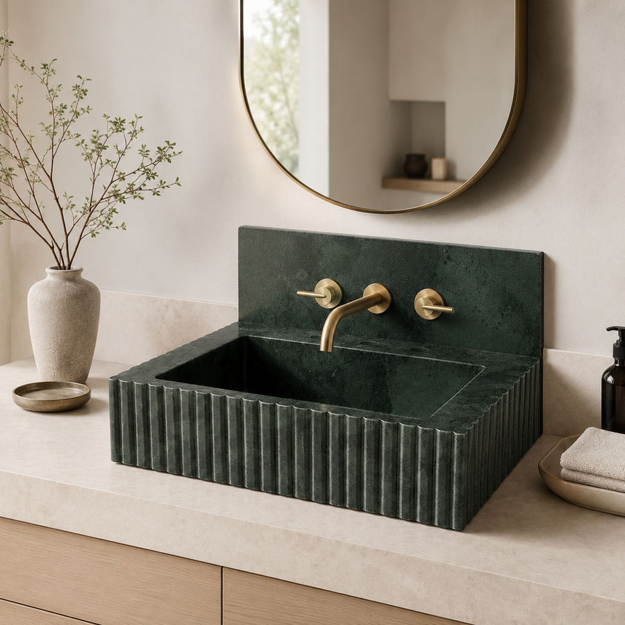 A dark green marble bathroom sink with a fluted front and a matching backplate, paired with gold-tone faucets, set on a light beige countertop in a styled bathroom scene.