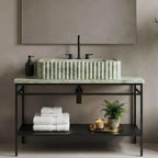 A modern bathroom vanity setup with a rectangular green marble vessel sink on a ribbed green marble countertop, mounted over a black metal console table. White folded towels, a black pump bottle, a gold tray with a candle, and a potted fern sit below, against a warm taupe wall with a large mirror above.