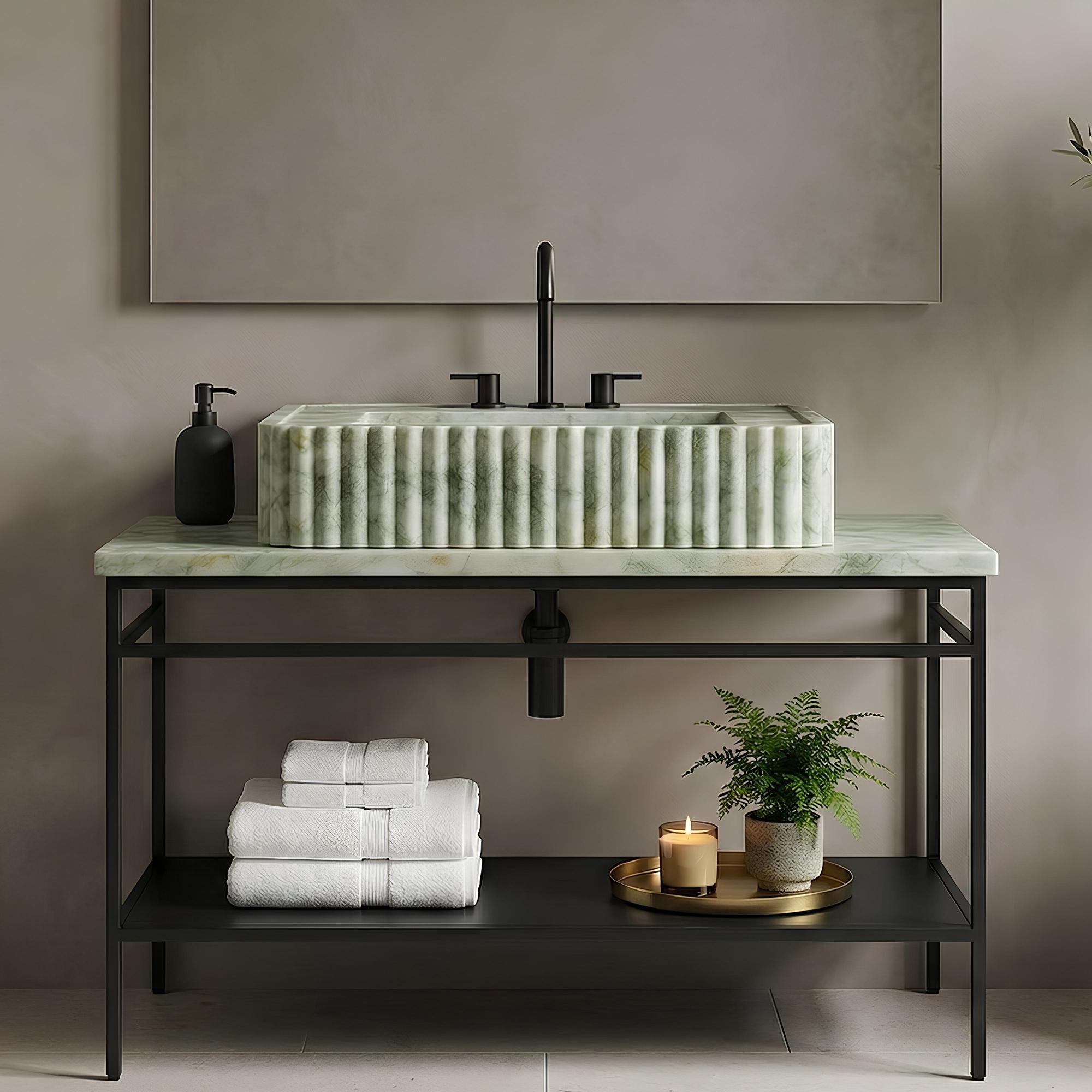 A modern bathroom vanity setup with a rectangular green marble vessel sink on a ribbed green marble countertop, mounted over a black metal console table. White folded towels, a black pump bottle, a gold tray with a candle, and a potted fern sit below, against a warm taupe wall with a large mirror above.