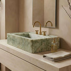 A rectangular green marble vessel sink with fluted sides, installed on a light stone countertop with a gold faucet, set in a warm beige bathroom.