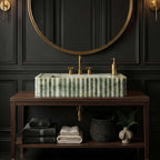 A rectangular marble bathroom sink with a ribbed fluted surface in green and white, mounted on a dark wood console table. The sink is styled with gold fixtures, including a matching gold faucet and soap dispenser, set against a dark paneled wall with a large round gold-framed mirror.