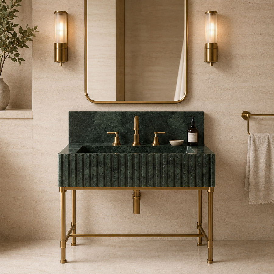 A modern bathroom vanity featuring a ribbed dark green marble sink and matching backplate with a brushed brass faucet, handles, and stand. The vanity is styled in a warm beige bathroom with a gold-framed mirror, two lit wall sconces, and a towel on a rack.