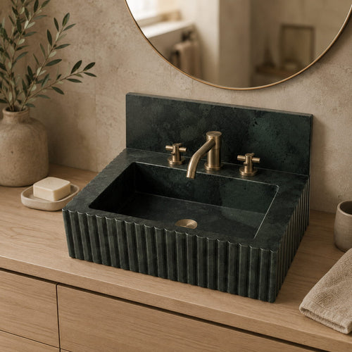 A dark green fluted stone bathroom sink with a matching backplate and brass faucet, installed on a light wood vanity with a round mirror and toiletry accessories in a softly styled bathroom setting.
