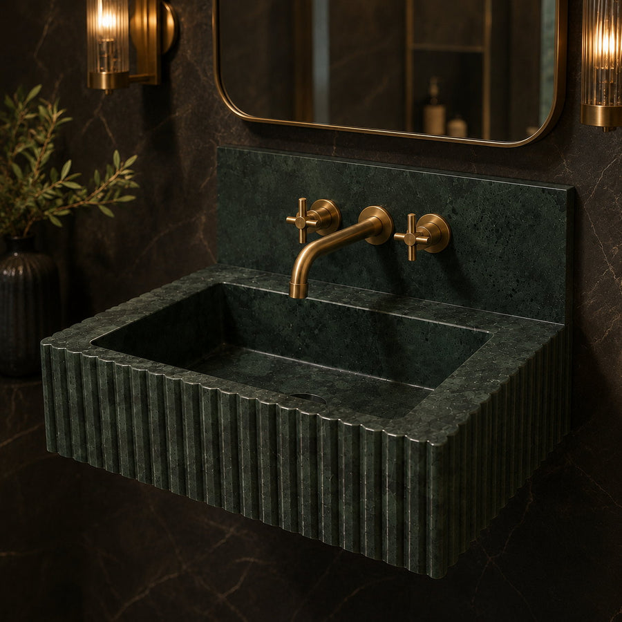 A modern floating bathroom sink made of dark green fluted stone with a matching rectangular backsplash, paired with a curved matte brass faucet and two cross-handle valves. The sink is mounted on a dark marble wall beneath a gold-framed mirror, flanked by warm wall sconces.