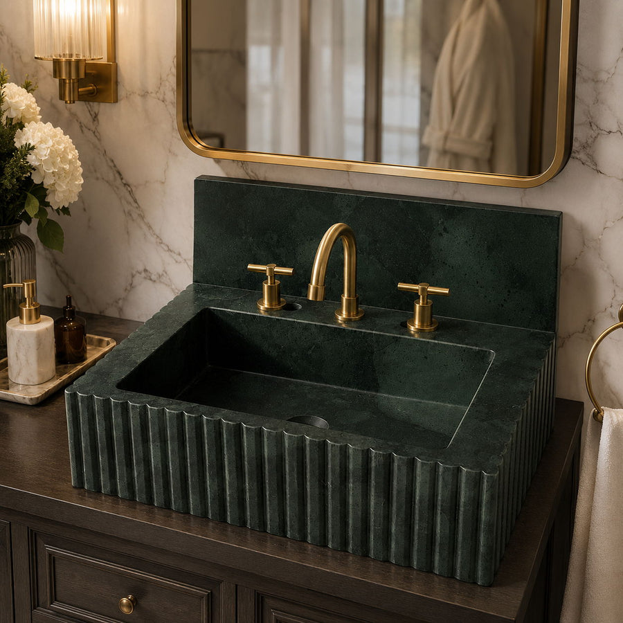 A dark green fluted marble bathroom sink with a matching backplate and gold faucet fixtures, installed on a dark wood vanity in a styled bathroom setting with a mirror, wall sconce, and flowers.