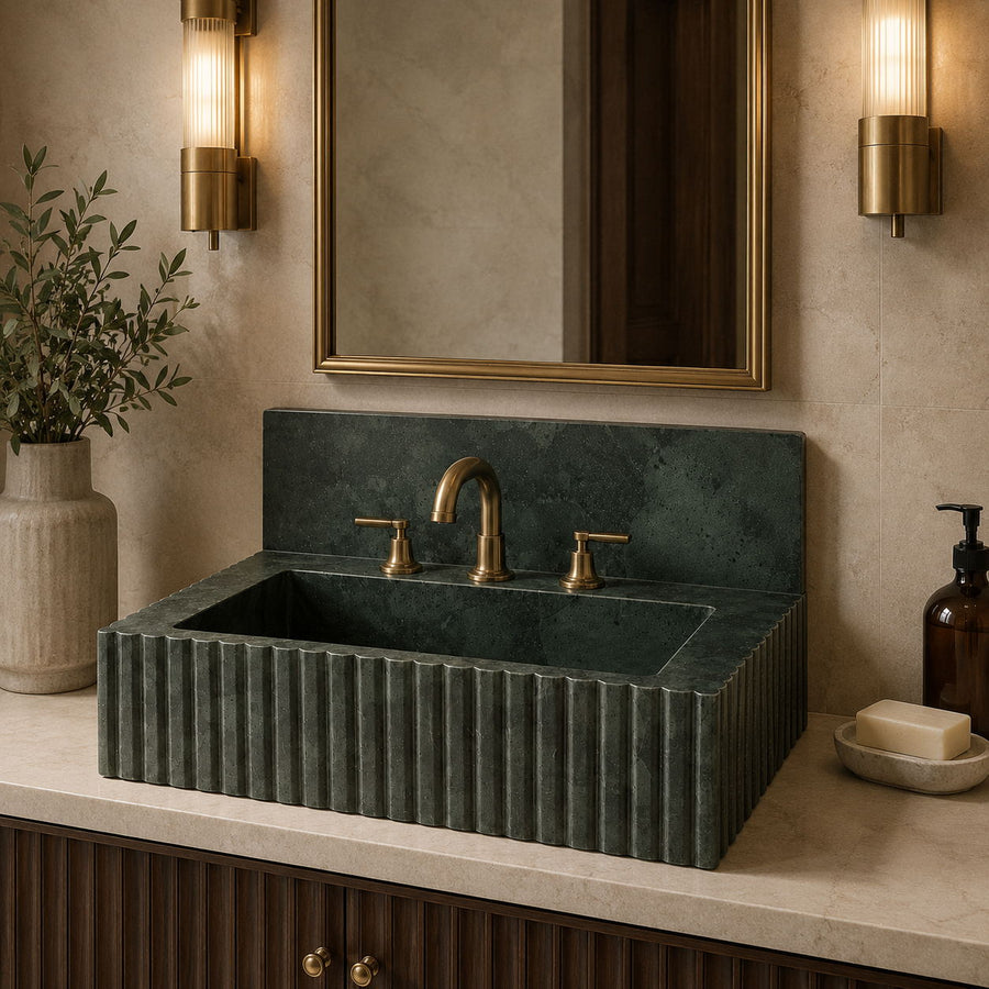 A dark green fluted stone vessel sink with a matching backplate and brass faucet, installed on a beige marble countertop in a bathroom setting with a gold-framed mirror and wall sconces.