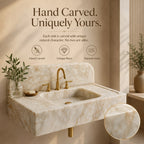 A wall-mounted marble sink with a gold faucet set, shown in a warm beige interior setting with decorative plants and text describing the hand-carved, unique piece nature of the sink.