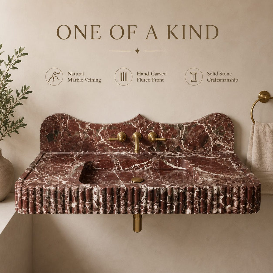 A freestanding rectangular sink with a dark red and white veined marble countertop and backsplash, paired with brass fixtures, shown against a beige wall with decorative text and a plant.
