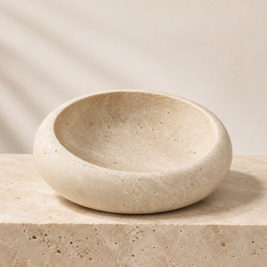 A round travertine stone soap dish with a textured natural stone finish, sitting on a matching stone pedestal against a neutral beige background.