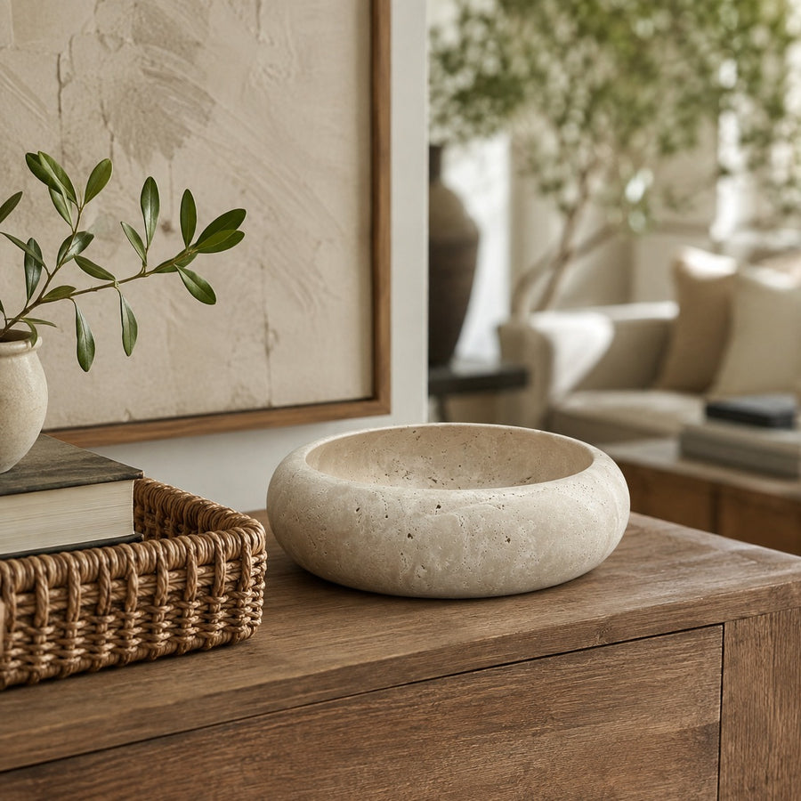 A round, shallow stone bowl in a warm beige color sits on a wooden console table in a styled living room setting.