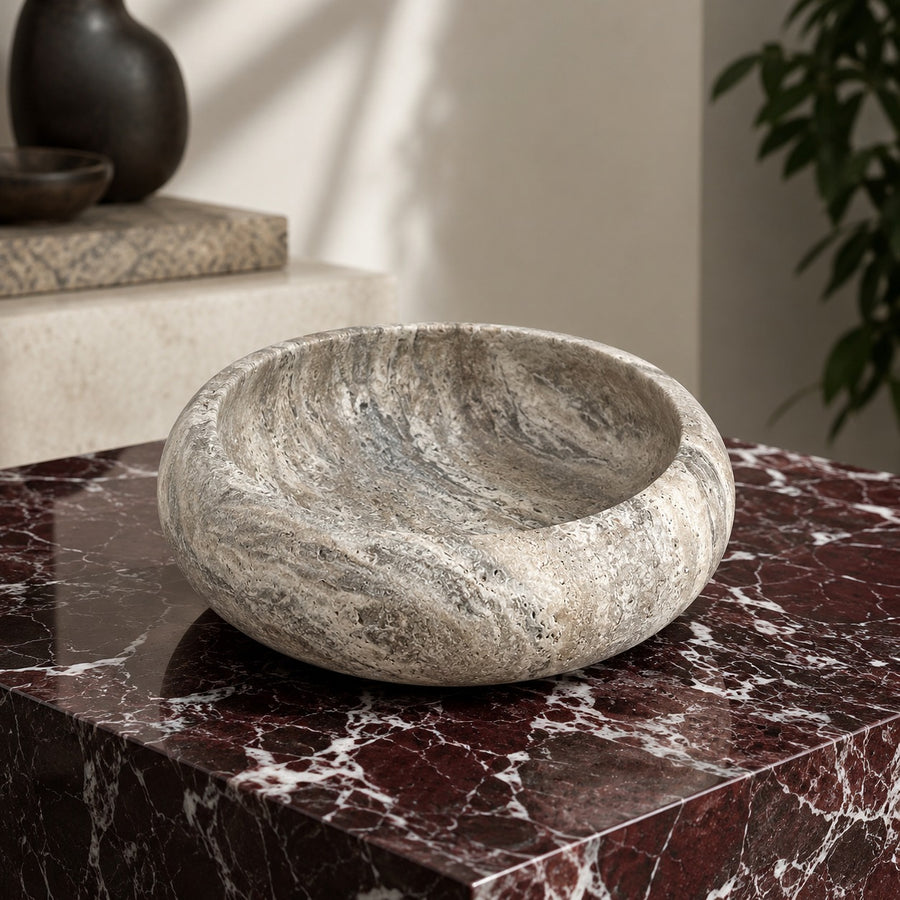 A round stone vessel sink or decorative bowl made of textured gray and beige marble, shown resting on a dark red marble surface in an interior setting.