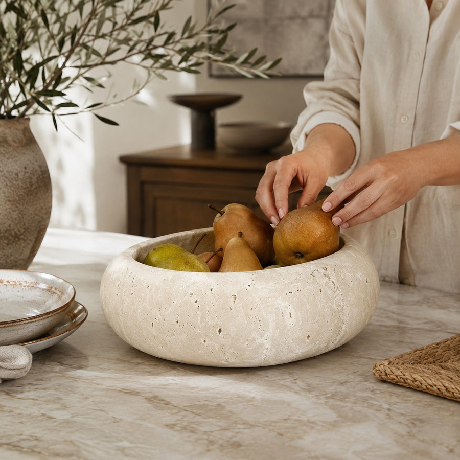 A round, textured stone fruit bowl filled with pears on a marble table, with a person's hands and torso visible adjusting the fruit. The scene is styled in a warm, modern kitchen or dining setting.