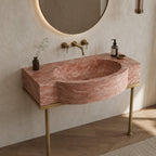 A modern wall-mounted bathroom sink with a pink marble-look basin and gold metal legs, shown installed against a textured beige wall with a round mirror, brass faucet, and small decorative bottles.