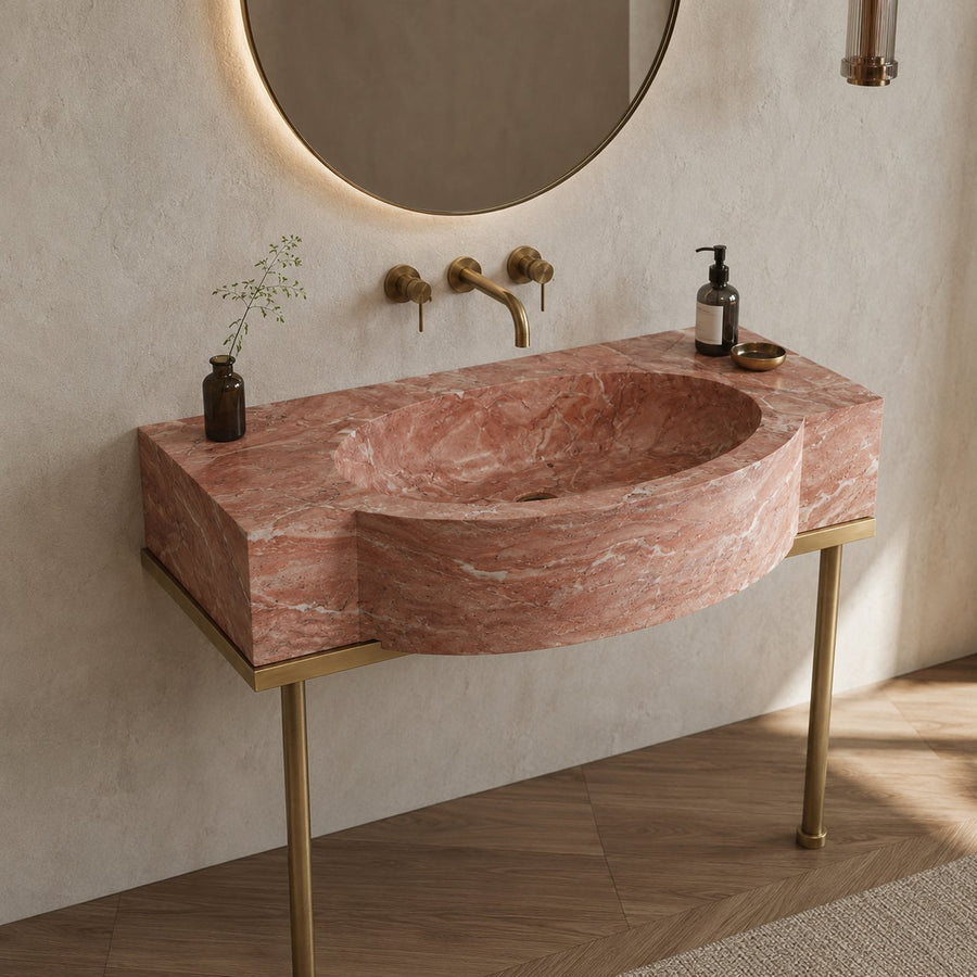 A modern wall-mounted bathroom sink with a pink marble-look basin and gold metal legs, shown installed against a textured beige wall with a round mirror, brass faucet, and small decorative bottles.