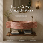A round vessel sink made of pinkish-red natural marble with white veining, mounted on a wooden floating vanity with brass legs. The scene is styled like a bathroom with a round mirror, a lit wall sconce, a vase with greenery, and folded towels on the shelf.
