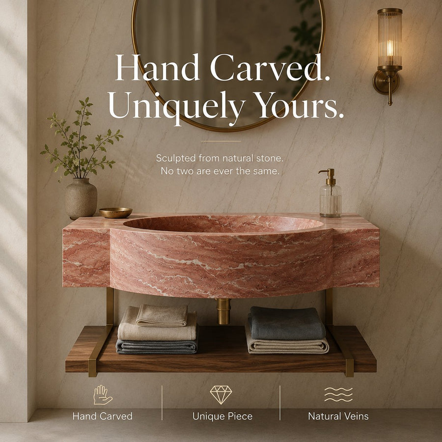 A round vessel sink made of pinkish-red natural marble with white veining, mounted on a wooden floating vanity with brass legs. The scene is styled like a bathroom with a round mirror, a lit wall sconce, a vase with greenery, and folded towels on the shelf.