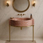 A modern bathroom vanity featuring a curved pink marble sink with a matching marble countertop, supported by elegant gold-toned metal legs. The vanity is styled with a round gold-framed mirror, wall-mounted brass sconces, a gold faucet set, and a beige woven rug on a light marble tiled floor.
