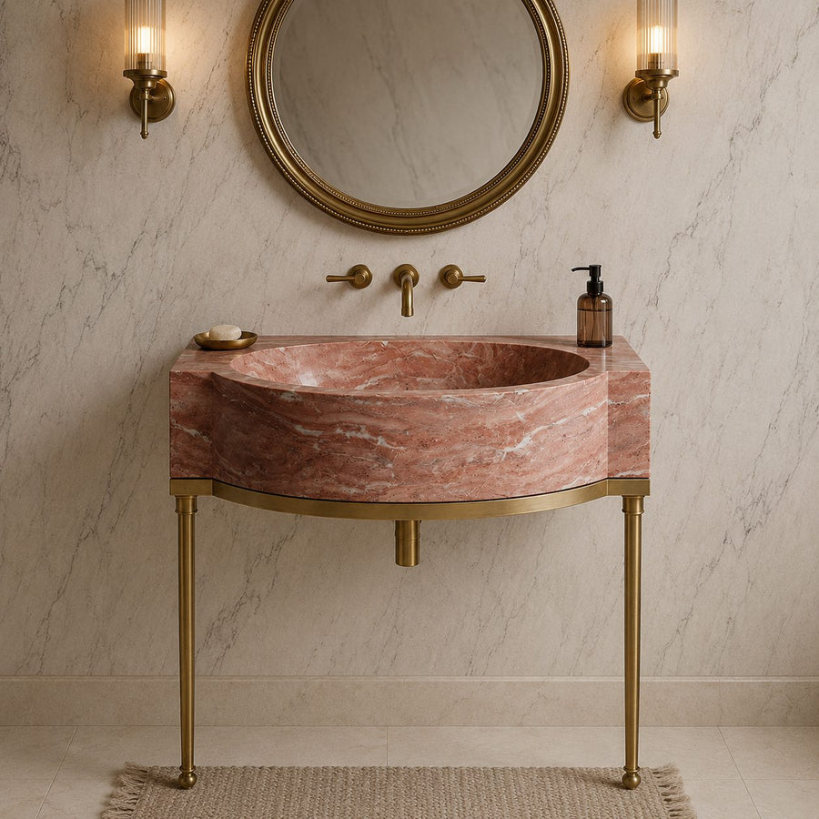 A modern bathroom vanity featuring a curved pink marble sink with a matching marble countertop, supported by elegant gold-toned metal legs. The vanity is styled with a round gold-framed mirror, wall-mounted brass sconces, a gold faucet set, and a beige woven rug on a light marble tiled floor.