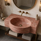 A round vessel sink made of pinkish-red marble with natural veining, mounted on a matching marble countertop with brass fixtures, set in a modern bathroom.